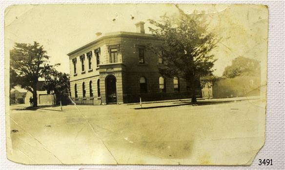 Sepia photograph of two storey building on street corner. Figures under tree on left.