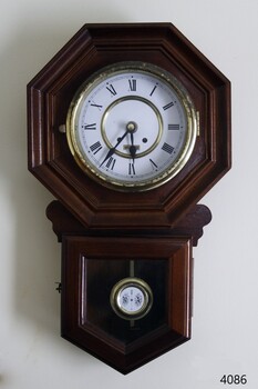 Dark wood case, octagonal shape around clock face, decorative pendulum, Roman numerals on dial.