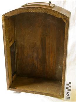 Open fronted wooden clock case on bench. Top is curved. There is a door at the top and on the side. 