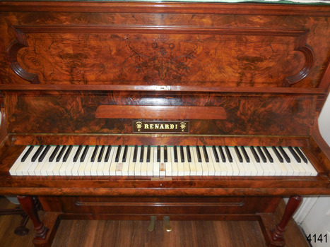 Complete width of the keyboard and the lock in the centre, below the keyboard. The music ledge has curved edges. The lid has a brass hinge from left to right along the join. The two brass pedals can be seen.