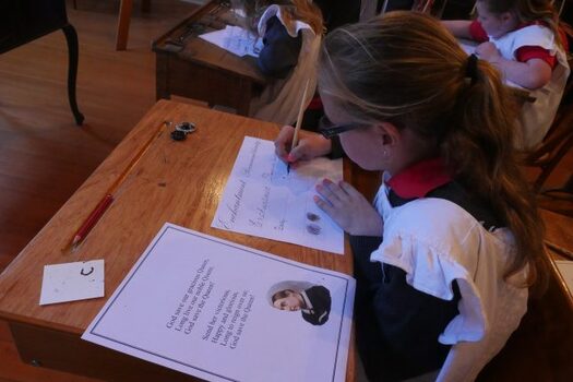 Colour photograph showing re-enactment of a schoolgirl in old fashioned dress writing at her desk. 
