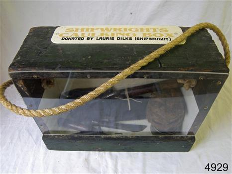 Wooden toolbox has a Perspex front and contains objects for caulking. A sign is attached to the top.