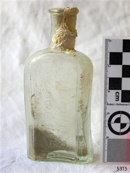Clear glass bottle. Contains sediment. White encrustations on neck.