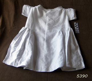 White dress has plain bodice and tucks at the waist. Lade trim is added to neckline and short sleeves.
