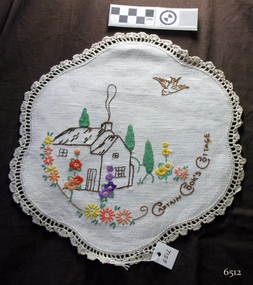 Ovate doily of white cotton with embroidered cottage scene and crocheted edging