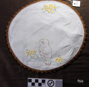 Round white doily with embroidered kookaburra on branch, wattle flowers and leaves, and brown crocheted edge