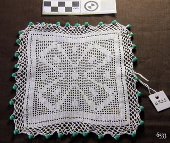 White square crocheted jug cover with a Maltese Cross incorporated in the design and a border of green beads