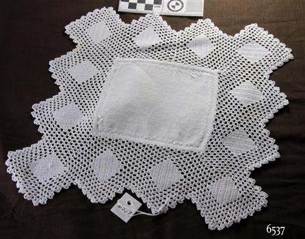 Overall square white crocheted doily with diamond patterns, with rectangle of white linen inserted in centre and decorated with cross stitch