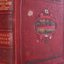 Large red bound book, gold writing and image of a ship etched on the cover. 