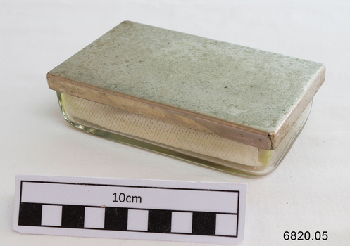 A small rectangular glass container with a metal lid containing lint, angle-ended tweezers, glass syringe and a separate metal end piece.