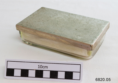 A small rectangular glass container with a metal lid containing lint, angle-ended tweezers, glass syringe and a separate metal end piece.