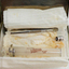 A small rectangular glass container with a metal lid containing lint, angle-ended tweezers, glass syringe and a separate metal end piece.