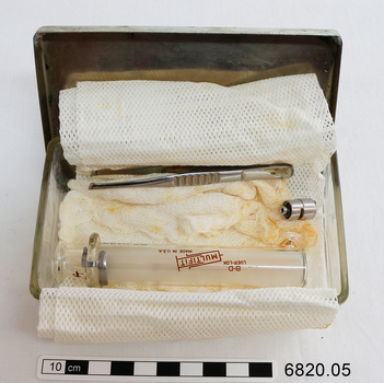 A small rectangular glass container with a metal lid containing lint, angle-ended tweezers, glass syringe and a separate metal end piece.