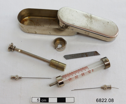 An oblong stainless steel metal box with lid, containing items comprising a syringe set, namely needles, plunger, cylinder, blade, etc.