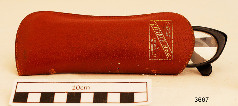 Plastic rimmed reading spectacles in a tan case with printing in gold on the outside showing the maker's name and address.