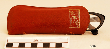 Plastic rimmed reading spectacles in a tan case with printing in gold on the outside showing the maker's name and address.