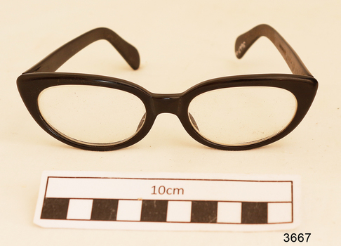 Black plastic rimmed reading spectacles in a tan case with printing in gold on the outside showing the maker's name and address.