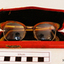 Orange plastic rimmed reading spectacles in an red case with printing in gold on the outside showing the maker's name and address. Part of the plastic frame is broken. Hinges of spectacles have seized up.