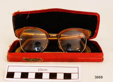 Orange plastic rimmed reading spectacles in an red case with printing in gold on the outside showing the maker's name and address. Part of the plastic frame is broken. Hinges of spectacles have seized up.