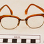 Orange plastic rimmed reading spectacles in an red case with printing in gold on the outside showing the maker's name and address. Part of the plastic frame is broken. Hinges of spectacles have seized up.