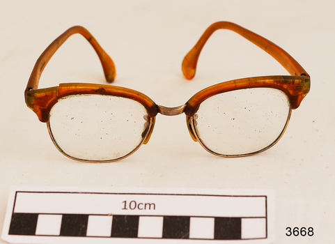 Orange plastic rimmed reading spectacles in an red case with printing in gold on the outside showing the maker's name and address. Part of the plastic frame is broken. Hinges of spectacles have seized up.