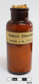 Brown glass bottle with thin lip, wide neck, rounded shoulders, body tapering inwards to a slightly narrower base. Cream coloured paper label just below shoulders has black print text. 