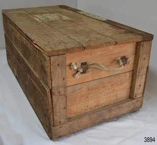 Oblong wooden trunk made from planks, with hinged lid on the long side. Rope handles on both short sides. Address of owner on lid.