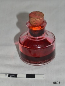 Round clear glass ink bottle with cork stopper. About one third of the bottle contains red ink.