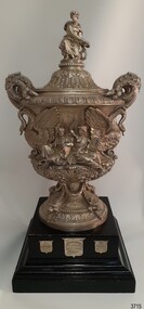 Elaborately decorated silver trophy with lid on a black plinth with award shields on it