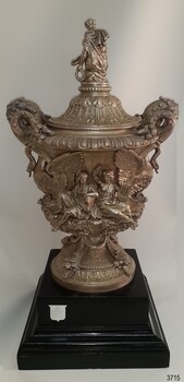 Award shield on black plinth. Back of figure on lid, sides of figures on the handles.