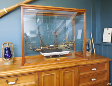 Ship model is in wooden framed glass-sided case, bow facing left.