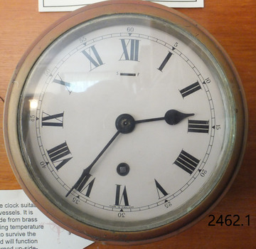 Brass rimmed, white faced clock with Roman numerals, mounted on a board.