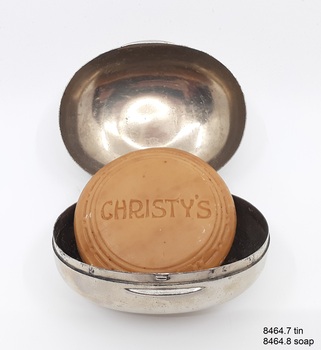 Oval metal box with hinged lid containing a complete cake of soap