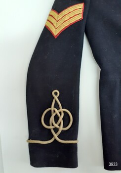 Right sleeve has Sergeant stripes (three chevrons)  at shoulder and gold braid fobbing above cuff