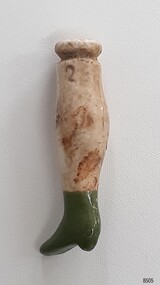 Cream coloured ceramic doll's leg with green heeled boot and groove around flat round top. It has an inscription.