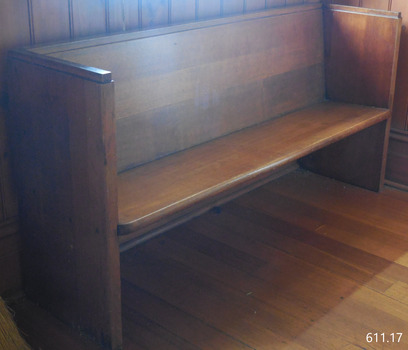 The pew is inside the doorway facing into the school room. It is along the east wall against the south wall.