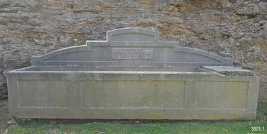 Rectangular concrete horse trough with arched pediment or panel positioned across the back. In the centre of the pediment is an inscribed plaque. The front of the trough has a design of three panels across its width, and these line up with the horizontal shoulder of the pediment. 
