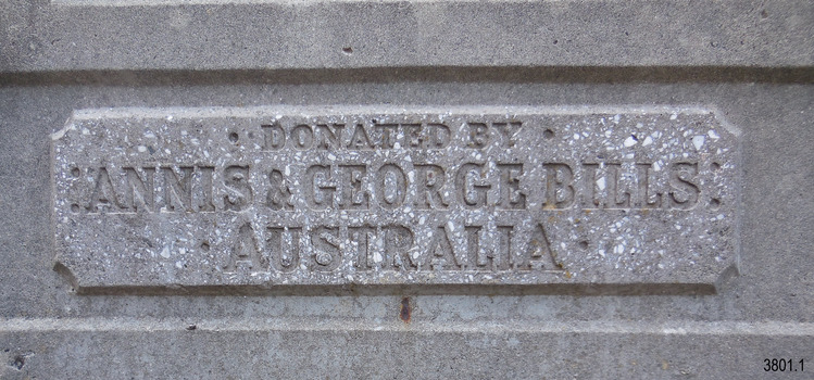 The inscription has been moulded into a bevelled, rectangular plaque on the concrete pediment. The text is all in capital letters spread across three rows. The top row is in smaller letters. The words are “Donated by Annis and George Bills Australia”.