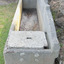 The trough contains a little water that has gathered at the left end. The cap is not quite fitted over the compartment, reversing the built-in ledge where the cap would rest in place. On the closest end there are four remnants of metal, spaced like the corners of a rectangle and within lighter coloured, round concrete areas, suggesting possible removal of a fitting, perhaps with a grinder. Towards the bottom of this end is another round, lighter coloured area. The end of the pediment rests on the top of the right end. The different colours of the inside of the trough are visible.