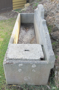 The trough contains a little water that has gathered at the left end. The cap is not quite fitted over the compartment, reversing the built-in ledge where the cap would rest in place. On the closest end there are four remnants of metal, spaced like the corners of a rectangle and within lighter coloured, round concrete areas, suggesting possible removal of a fitting, perhaps with a grinder. Towards the bottom of this end is another round, lighter coloured area. The end of the pediment rests on the top of the right end. The different colours of the inside of the trough are visible.
