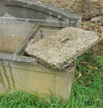 The cap is resting at a 45 degree angle across the top of the compartment. Reinforcing shows at the front right corner where the concrete has been chipped off. The left and rear edges of the ledge for the cap are visible.