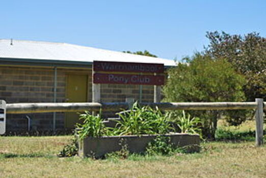 Horse trough is in front of the Warrnambool Pony Club sign. There is a grey brick building in the background. The trough is filled with green leafy plants.  The photograph was published in Wikimedia Commons, on a page called Bills Horse Troughs.