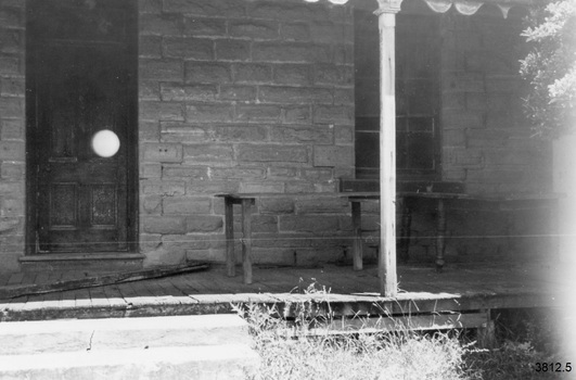 Front right of building. On the left is a panelled door, on the right a multi paned window. The border of stones around the window seems to be a similar design as the other windows of the building but these border stones seem all the same colour as the other wall stones on this wall. There are short timber tables or benches on the veranda, some have turned legs. There are rotted timber boards laying on the veranda. The veranda post is square and has a decorative top. Above this, along the veranda roof, is a scalloped pattern; perhaps metal. Steps have been placed in front of the veranda to line up with the door. There are leaves on branches on the right. 
