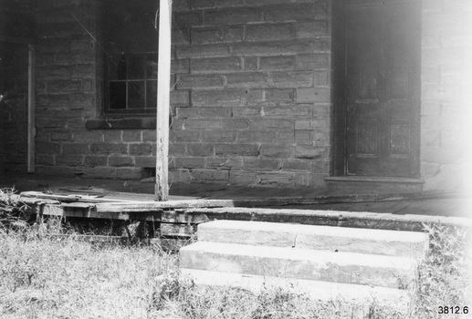 Front left of building. The steps and timber on the veranda line up with view 5. On the left there is a post and beam attached to the stone wall. There is a 12-paned window near the centre and a panelled door on the right. Below the window at floor level there is a space where a stone has been removed or broken away. On the veranda in front of the door is a rotting piece of timber. The veranda floor boards are detreating and breaking away. A square veranda post has uncoiled wire or twine handing beside it. In front of the veranda is a set of three steps, placed to lead to the door.