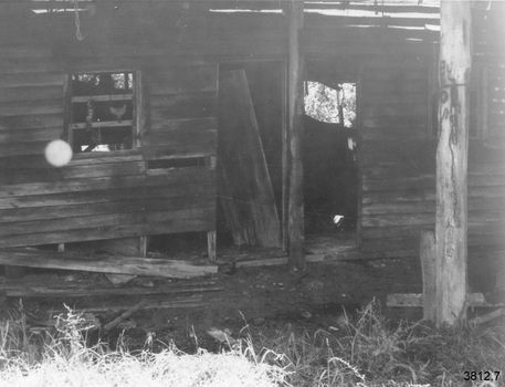 Timber outbuilding. Timber planks have fallen off the walls. In the centre are two doorways beside each other, the left doorway has a door resting across it. On the left of this doorway is a window without glass. On the right of the right doorway is another window. Light is showing from the other side of the building. On the right, a short distance from the wall, there is a deteriorating post.