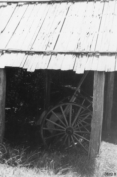 Covered shelter containing a two-wheeled light-weight cart. The roof is peaked and made from timber boards, roughly trimmed. Four chunky square timber posts support the roof. Trees or bushes appear to be on the other side of the area and the ground is grassed. The cart's wheels have 16 spokes, which seem to be timber, and what appears to be a metal rim and hub.