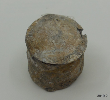 Black-grey lead seal has encrustations, top is separated from sides and is very thin.