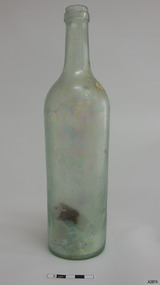 Tall, clear glass bottle. Shoulder seam. Cork has fallen into base. Glass shows opalescence. Encrustation on surface.