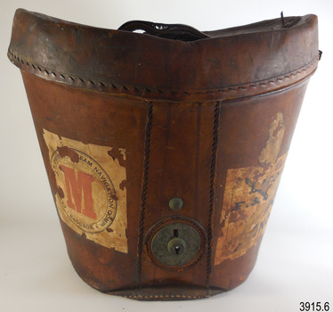 Brown leather hat box with curved lid. It has a carry handle and metal lock. Labels are attached.
