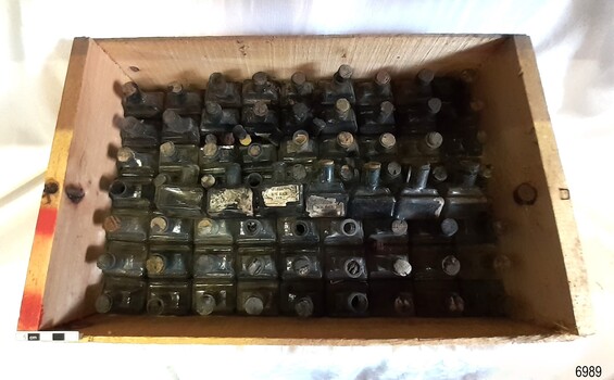 Rows of small glass inkbottles in a wooden crate, some wit corks